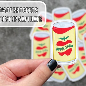 May include: A hand holds an "Apple Sidra" sticker, a cartoon design of a yellow can with a red apple graphic. The text on the sticker reads "Apple Sidra." Above the sticker is the text "50% of proceeds go to stop AAPI hate."