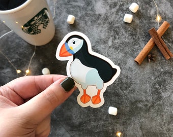 May include: A sticker of a cartoon puffin with a black and white body, orange beak and feet, and blue eyes. The sticker is on a gray surface.