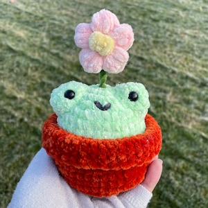 May include: A crocheted frog wearing a pot with a pink flower on top. The frog is green and the pot is orange.