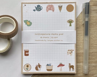 May include: A cottagecore themed memo pad with 48 sheets and 90 gsm paper. The pad features illustrations of a chicken, teapot, sweater, flowers, pie, duckling, hat, jar, sunflower, rabbit, yarn, mushroom, and deer. The text on the pad reads "cottagecore memo pad 48 sheets | 90 gsm handmade by lorynn sticker co."