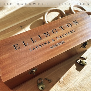 May include: A rectangular wooden box from the Exotic Hardwood Collection, featuring the engraved text ELLINGTON, SABRINA & ZACHARY, and 4/25/2026. The box has a metal latch, and small brass-colored padlocks and keys are placed around it.