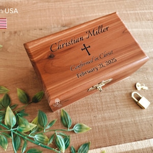 May include: A wooden box with a hinged lid, featuring the engraved name "Christian Miller", a cross, and the text "Confirmed in Christ February 21, 2025". The box has a brass clasp and lock. Green leaves are in the foreground.