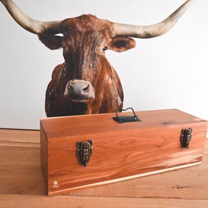May include: A rectangular wooden storage box with a reddish-brown grain, featuring two dark metal front latches and a black metal handle on the top lid. The box has a small logo printed in the bottom left corner on the front panel.