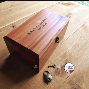 May include: A rectangular wooden keepsake box with a hinged lid and a metal latch. The lid is engraved with the text KATE & HENRY EST. 2026. A small metal padlock and a separate key are placed near the box. The wood has a natural grain.