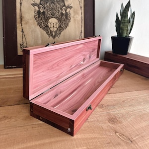 May include: Open wooden box with a pinkish-red interior and exterior. The box has a rectangular shape with a hinged lid and a metal clasp. The wood grain is visible, and the box is sitting on a wooden surface.