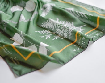 May include: Green silk scarf featuring a white botanical print of leaves and branches. The scarf has a gold and black border with the text "Leaves of Canada" and "The Flora of Canada".