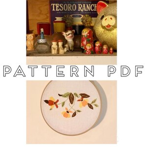 Może przedstawiać: Zbliżenie na gotowy tamborek z haftem kwiatowym. Wzór przedstawia trzy żółte i pomarańczowe kwiaty z brązowymi liśćmi i łodygami na białym tle z tkaniny. Tamborek jest wykonany z drewna i ma naturalne wykończenie.