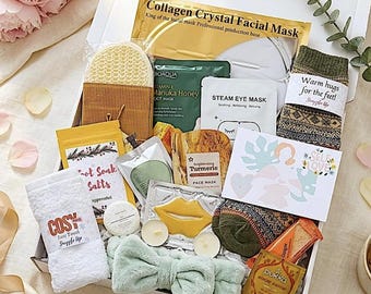 May include: A gift box filled with self-care items, including face masks, socks, a headband, and bath salts. The box contains a variety of products, such as a collagen facial mask, a steam eye mask, and a turmeric face mask. The box is decorated with a bow and a card that says "Self Love".