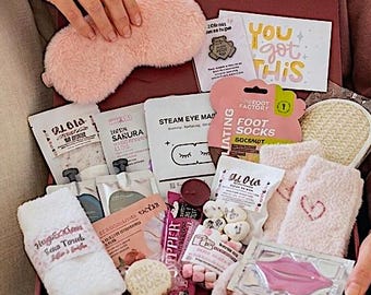 Könnte beinhalten: Eine bordeauxfarbene Geschenkbox gefüllt mit Pflegeprodukten, darunter eine flauschige rosa Augenmaske, kleine Handtücher und verschiedene Hautpflegeprodukte. Die Box enthält Artikel mit dem Text "YOU got THIS" und "FOOT SOCKS".