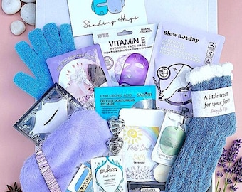 May include: A gift box filled with self-care items, including a blue glove, face masks, eye mask, socks, and tea. The box contains a card that says "Sending Hugs". The items are arranged on a pink surface with lavender and rose petals.