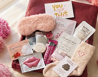 Può includere: Una confezione regalo per la festa della mamma con una mascherina per dormire rosa, maschere per il viso e calzini morbidi con un cuore. La scatola è circondata da fiori rosa e un'etichetta con la scritta "Happy Mother's Day".