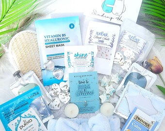 May include: A gift box filled with self-care items. Includes a sheet mask, bath salts, eye mask, headband, candles, and a bracelet. The box also contains a card that says "Hug in a Box" and "Sending Hu". The items are in shades of blue and white.
