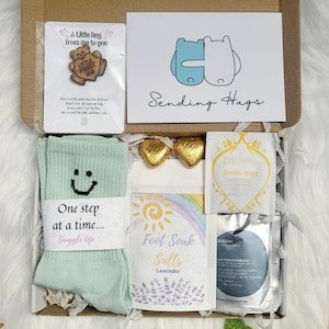 May include: A gift box containing self-care products. It features a card with the words "Sending Hugs", mint green socks with a smiley face and the text "One step at a time... Snuggle Up", lavender foot soak salts, and other items.