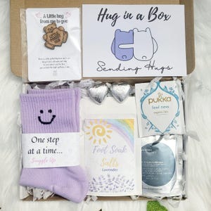 May include: A 'Hug in a Box' gift set containing lavender socks, foot soak salts, tea, and heart-shaped chocolates. The box also includes a card with the text "Hug in a Box" and a small pin.