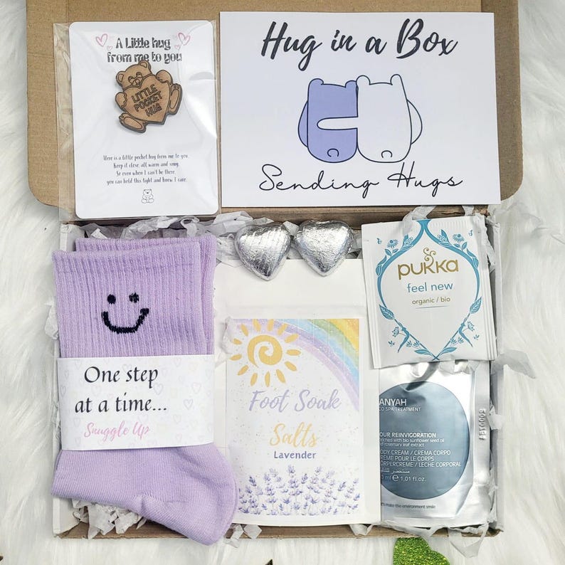 May include: A 'Hug in a Box' gift set containing lavender socks, foot soak salts, tea, and heart-shaped chocolates. The box also includes a card with the text "Hug in a Box" and a small pin.