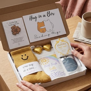 May include: An open gift box labelled "Hug in a Box" with a yellow sock, chocolates, tea, and foot soak salts. The box also contains a pin and a card with the words "Sending Hugs". The box is on a wooden surface.