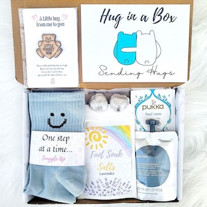 May include: A gift box filled with self-care products. It features a card with the text "A Little hug from me to you", blue socks with a smiley face, lavender foot soak salts, and other items. The box is labelled "Hug in a Box" and "Sending Hugs".