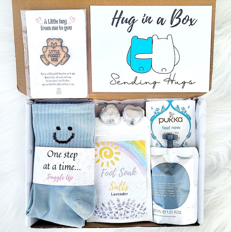May include: A gift box filled with self-care products. It features a card with the text "A Little hug from me to you", blue socks with a smiley face, lavender foot soak salts, and other items. The box is labelled "Hug in a Box" and "Sending Hugs".