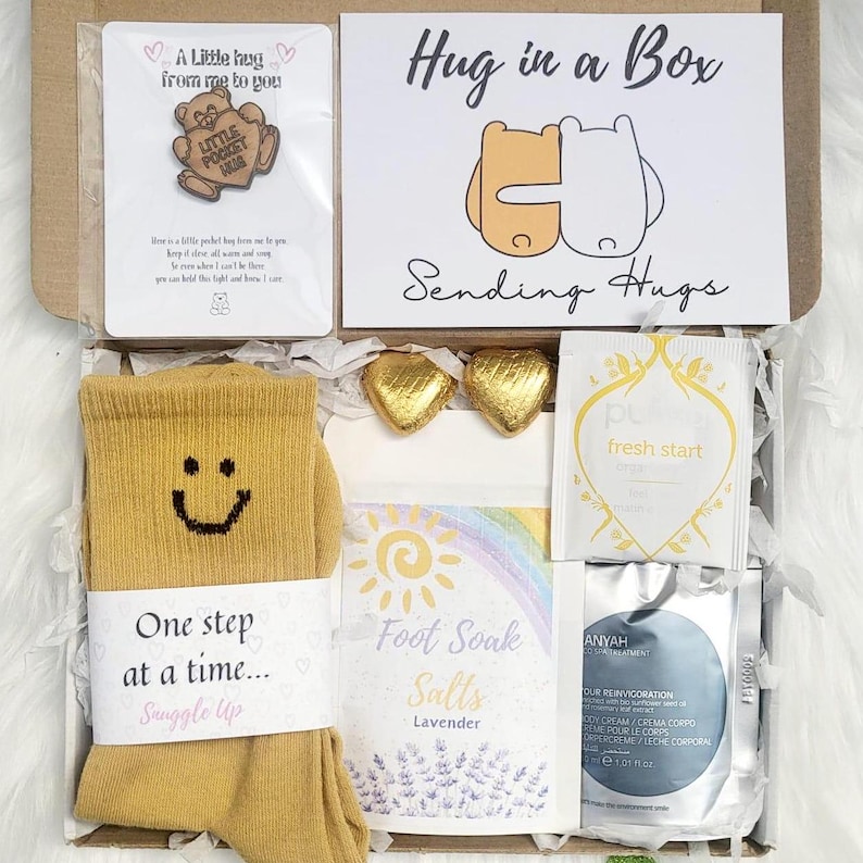 May include: A 'Hug in a Box' gift set. The box contains a pair of mustard yellow socks, a card with the text "Hug in a Box", lavender foot soak, and other items for relaxation and comfort.