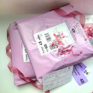 May include: Two pink packages with burgundy ribbon, shipping labels, and tags. One label reads "Tracked 48" and another says "Royal Mail Tracked". A small card reads "Share a Smile". Decorative pink paper is scattered around the packages.