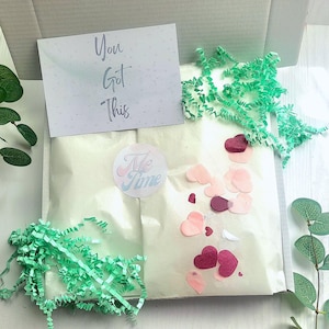 May include: A gift box with a white interior, containing a wrapped package, confetti, and a greeting card. The card reads "You Got This." A sticker with the words "Me Time" is also visible.