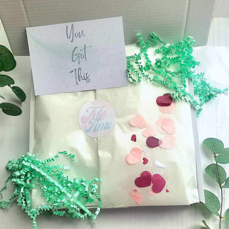 May include: A gift box with a white interior, containing a wrapped package, confetti, and a greeting card. The card reads "You Got This." A sticker with the words "Me Time" is also visible.