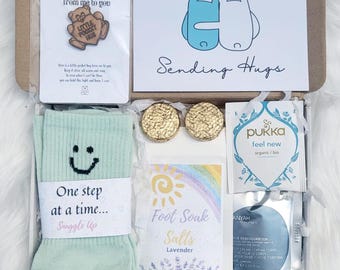 Può includere: Una confezione regalo piena di articoli per la cura di sé. Include un paio di calzini verde menta con una faccina sorridente, una carta con la scritta "Sending Hugs", una spilla con la scritta "Little Pocket Hug", sali da bagno, tè e due oggetti circolari color oro.