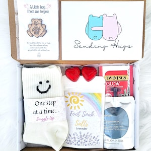 May include: A care package containing a variety of items. It features a card with a bear pin, a card with the words "Sending Hugs", socks, lavender foot soak salts, heart-shaped chocolates, tea, and body cream, all in a box.