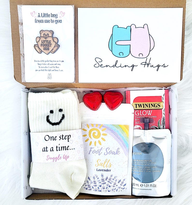 May include: A care package containing a variety of items. It features a card with a bear pin, a card with the words "Sending Hugs", socks, lavender foot soak salts, heart-shaped chocolates, tea, and body cream, all in a box.