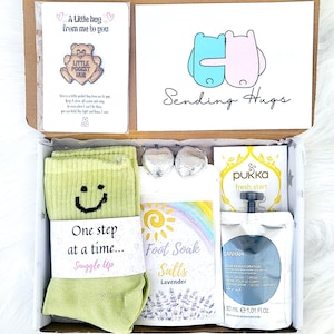 May include: A gift box containing self-care products. It features green socks with a smiley face, a card saying "Sending Hugs", lavender foot soak salts, a tea bag, and a body cream.