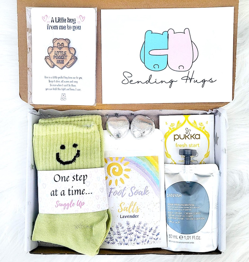 May include: A gift box containing self-care products. It features green socks with a smiley face, a card saying "Sending Hugs", lavender foot soak salts, a tea bag, and a body cream.