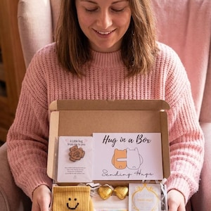 May include: A person holds a gift box labelled "Hug in a Box." The box contains yellow socks with a smiley face, a pin, chocolates, and other self-care items. The box includes the text "One step at a time..." and "Foot Soak Salts Lavender."