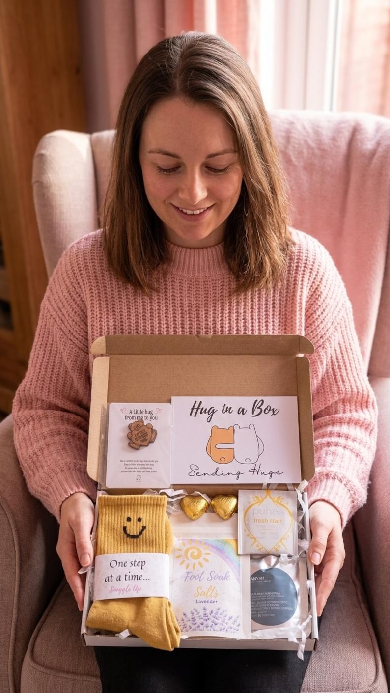 May include: A person holds a gift box labelled "Hug in a Box." The box contains yellow socks with a smiley face, a pin, chocolates, and other self-care items. The box includes the text "One step at a time..." and "Foot Soak Salts Lavender."