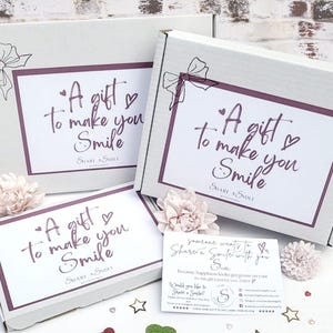 May include: Three white gift boxes, each with the message "A gift to make you Smile" in a handwritten style. The boxes are adorned with a bow and flowers. A small card with text is also present.