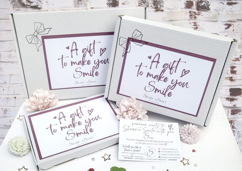 May include: Three white gift boxes, each with the message "A gift to make you Smile" in a handwritten style. The boxes are adorned with a bow and flowers. A small card with text is also present.