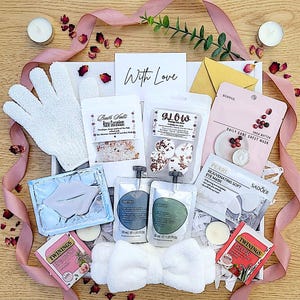 Op de afbeelding: Een spa-cadeauset met diverse zelfzorgproducten, waaronder badzout, gezichtsmaskers, oogmaskers en thee. De set is gerangschikt in een witte doos en versierd met een roze lint, rozenblaadjes en een kaart met "With Love".