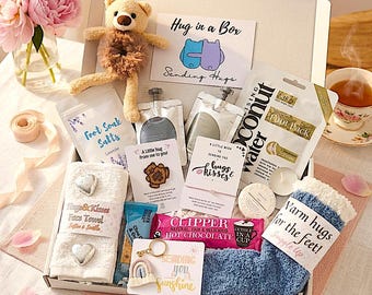 May include: A gift box containing a small teddy bear, lavender foot soak salts, coconut water foot pack, face towel with heart accents, Clipper hot chocolate, blue fuzzy socks, a rainbow keychain, and cards with messages like Hug in a Box.