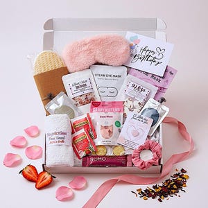Op de afbeelding: Een cadeaupakket met wellnessproducten, waaronder een roze oogmasker, een gezichtshanddoek, badzout, thee en een verjaardagskaart. De doos bevat ook een scrunchie, snoep en een chocoladereep.