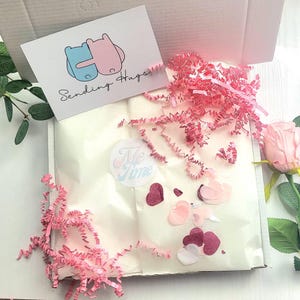 May include: A white box, open to reveal pink shredded paper, a card with two hugging bears and the words "Sending Hugs", and a sticker that says "Me Time". The box also contains pink and red heart-shaped confetti.
