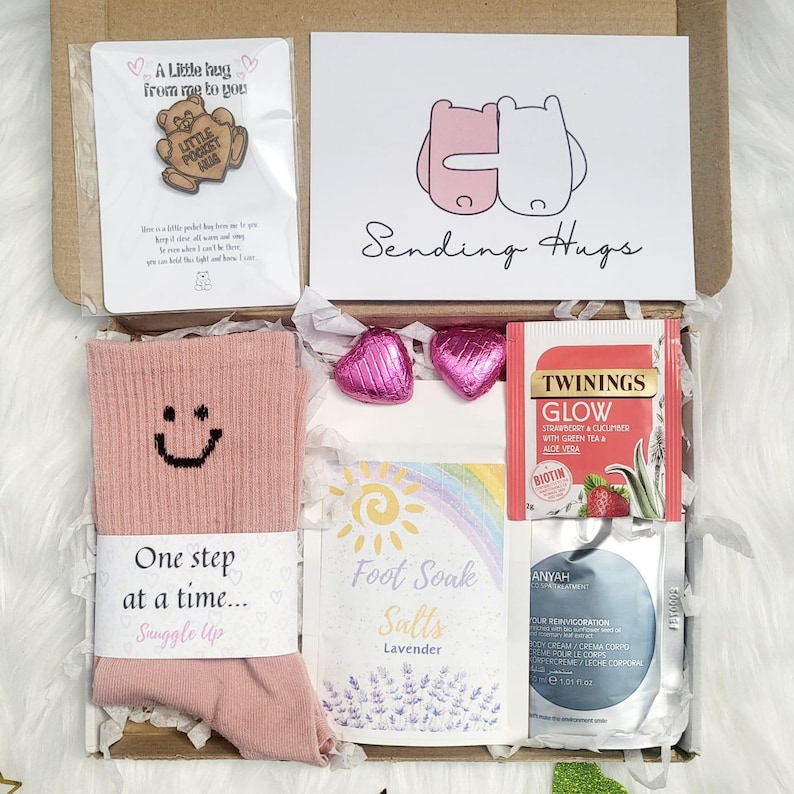 May include: A self-care gift box containing a pink sock with a smiley face, a card with the text "Sending Hugs", heart-shaped chocolates, tea, lavender foot soak salts, and a small pin.