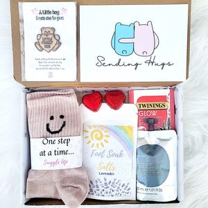 May include: A care package containing a pair of pink socks, lavender foot soak salts, heart-shaped chocolates, tea, and a card with the words "Sending Hugs". The box also includes a small pin.