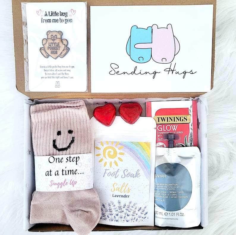 May include: A care package containing a pair of pink socks, lavender foot soak salts, heart-shaped chocolates, tea, and a card with the words "Sending Hugs". The box also includes a small pin.
