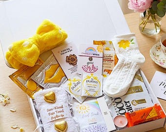 Può includere: Una confezione regalo bianca piena di articoli per la cura di sé. Include una fascia per capelli a fiocco gialla, maschere per il viso, calzini bianchi con un fiore giallo, un asciugamano bianco e altri prodotti di bellezza.
