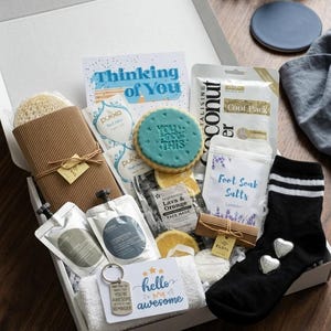 May include: A white gift box containing self-care products. Includes a card saying "Thinking of You", a cookie, face masks, foot soak salts, a loofah, a keychain, a towel, and black socks with heart details.