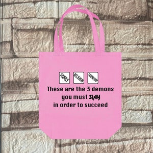 May include: Pink tote bag with a white graphic that says "These are the 3 demons you must slay in order to succeed." The graphic also includes three squares with the words "Family", "Religion", and "Friendship" inside.