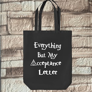 以下が含まれることがあります： 白い文字で「Everything But My Acceptance Letter」と書いてある黒いトートバッグ。中に円が描かれた白い三角形のシンボルがあります。