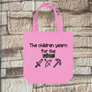 Puede incluir: Bolsa de tela rosa con el texto "The children yearn for the mines" e imágenes pixeladas de una espada, un arco y una flecha, y un pico.