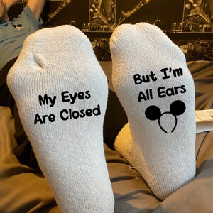 May include: A pair of white socks with black text. One sock says "My Eyes Are Closed" and the other says "But I'm All Ears" with a Mickey Mouse ear headband graphic.