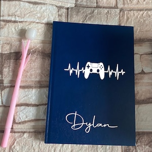 Op de afbeelding: Een donkerblauw notitieboek met een witte gamepad en een hartslaggrafiek. De naam "Dylan" is in wit cursief onder de grafiek geschreven.