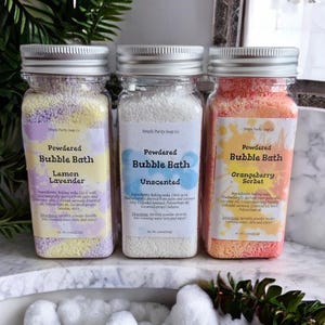 May include: Three glass jars filled with white, yellow, and pink bath salts. The jars have silver lids and labels that say "Powdered Bubble Bath" with different scents: Lemon Lavender, Unscented, and Orangeberry Sorbet.